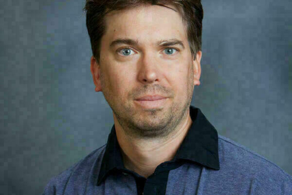 Headshot of a man with short brown hair and blue eyes. He wears a collared, short-sleeved, heather-blue shirt with a black accent on the collar. The background is gray.