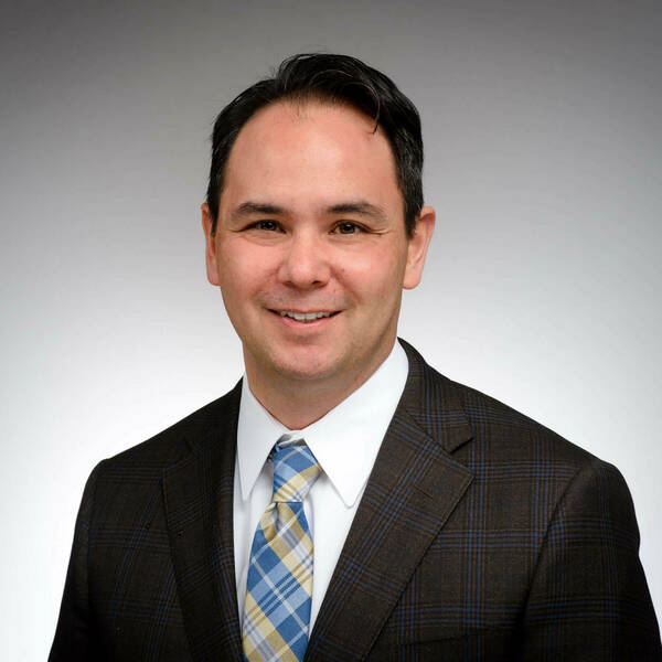 David Go, a smiling man with dark hair in a brown plaid suit with blue lines, white shirt, and blue-yellow plaid tie against a gray background.